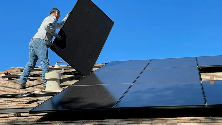 Installateur tilt een zwart zonnepaneel op een schuin dak onder een strakblauwe lucht